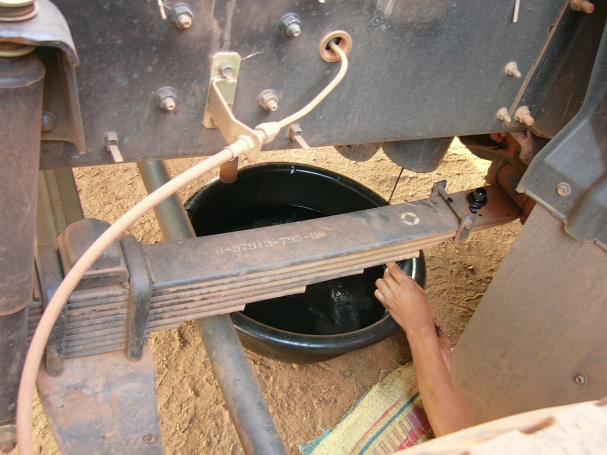 A leaf spring pack under a working truck in the field, covered in dust; a hand checks the area below the leaves